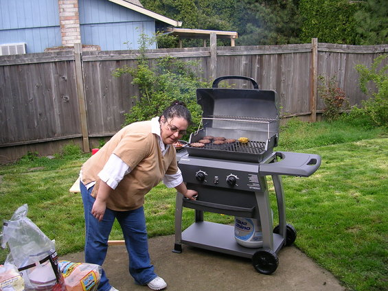 Woman in jeans and a tan shirt grilling hamburgers on a propane grill in a backyard