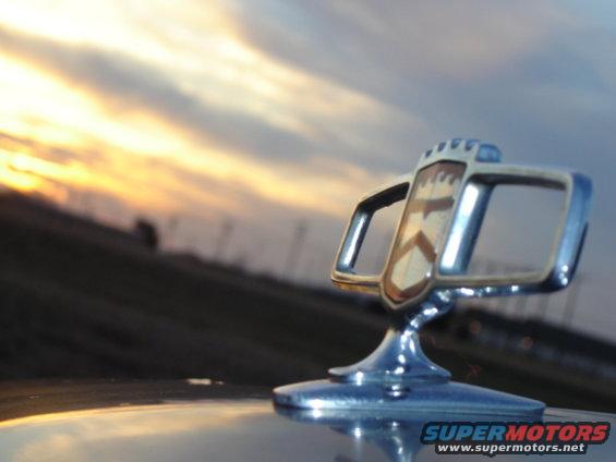 Chrome hood ornament with shield emblem against a sunset sky