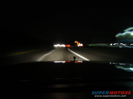 View from inside a car at night, looking at the dashboard and road ahead with headlights illuminating the way