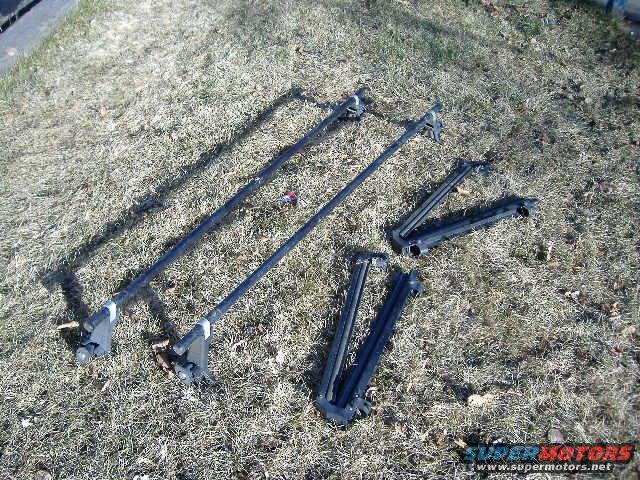 Two roof rack crossbars and four mounting feet laid out on dry grass
