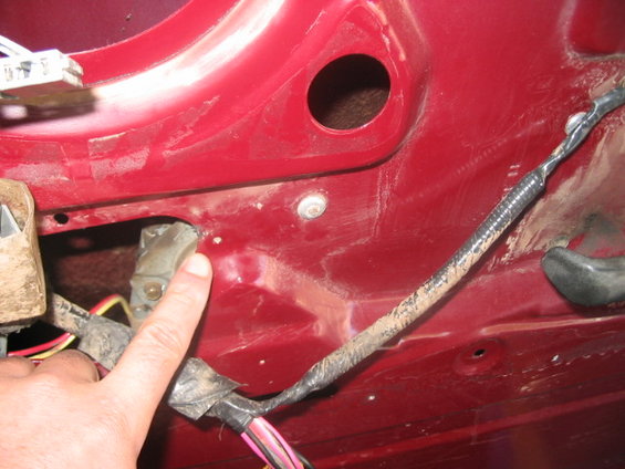 Close-up of red car door interior showing wiring harness and latch mechanism. Finger points to a component
