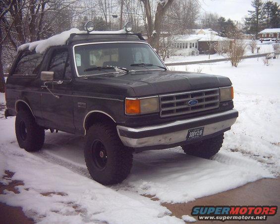 1987 Ford Bronco parked in snow, front 3/4 view. Has roof rack with lights and aftermarket wheels