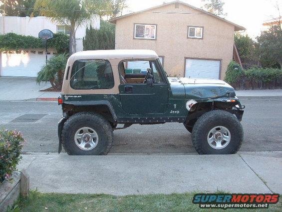 Green Jeep Wrangler parked on street, tan soft top, large tires, dented hood