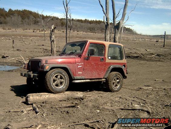 Red Jeep Wrangler with tan top parked on muddy ground with dead trees in background