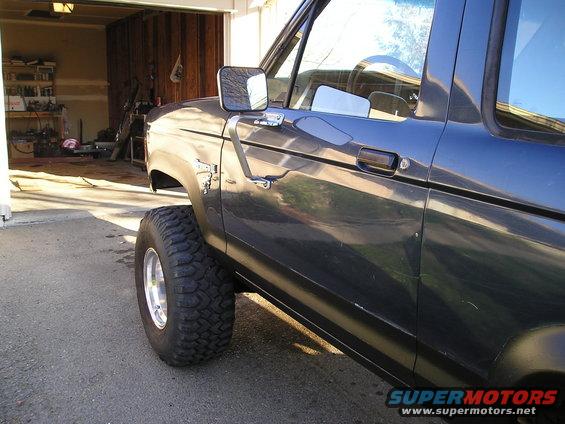Dark blue 1986 Ford Bronco II, side view, showing the front fender, door, and large off-road tire. Garage in background