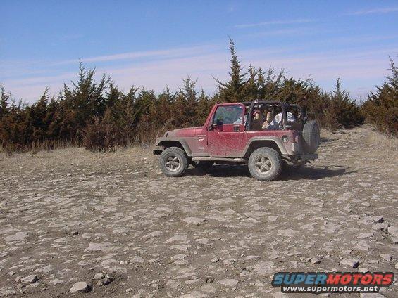 Red 1998 Jeep TJ driving on a rocky, unpaved trail with trees in the background