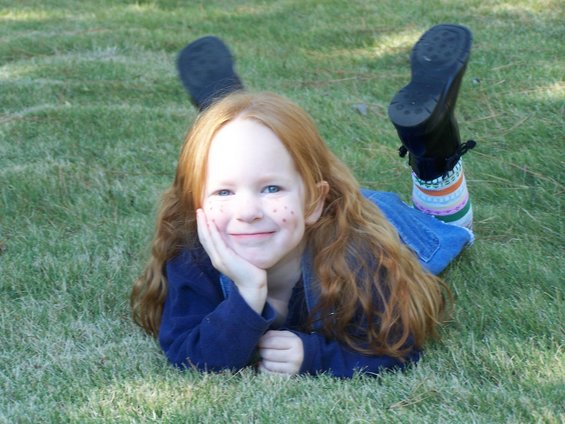 Young girl with long red hair and freckles lies on green grass, resting her chin on her hand