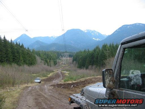 Jeep Wrangler in foreground, another SUV on muddy mountain road with power lines and mountains in background