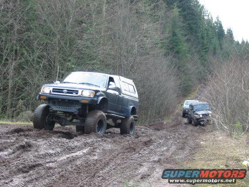 Modified Toyota pickup truck with large tires driving through mud, another SUV behind it on a wooded trail
