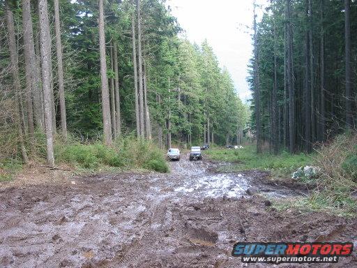 Muddy forest road with several off-road vehicles driving through it. SuperMotors logo at bottom right