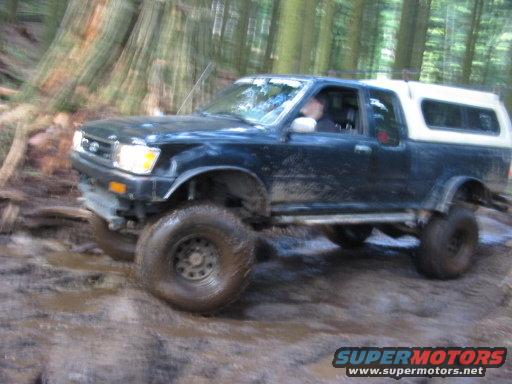 Dark blue pickup truck with white camper shell driving off-road through mud and trees