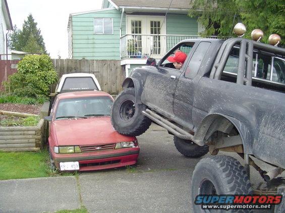 Red sedan with a white cap on its bed, parked on grass, with a black lifted pickup truck on top of its hood