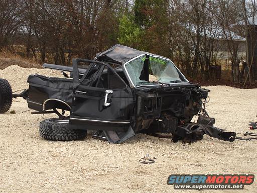 Wrecked black truck, front end missing, doors open, cracked windshield with text