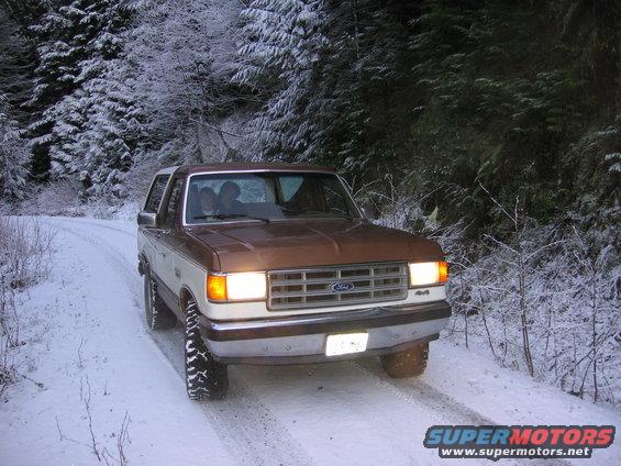 Brown and white 1987 Ford Bronco driving on a snowy road, front 3/4 view