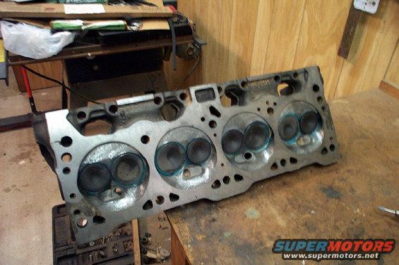 Engine cylinder head with four combustion chambers visible, resting on a workbench