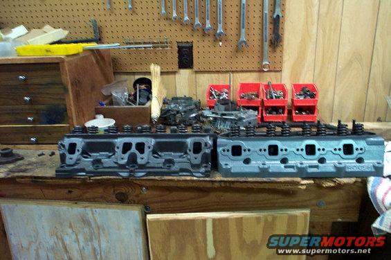 Two engine cylinder heads on a workbench, one gray, one blue with 'ROUSH' visible