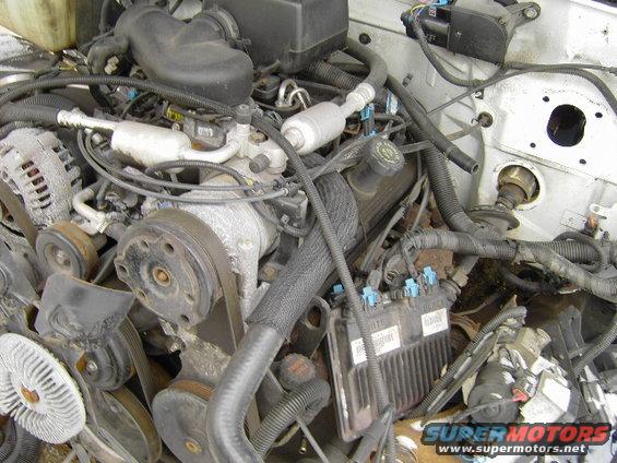 Engine bay with various hoses, belts, and electrical components visible. Some text is present on a black box