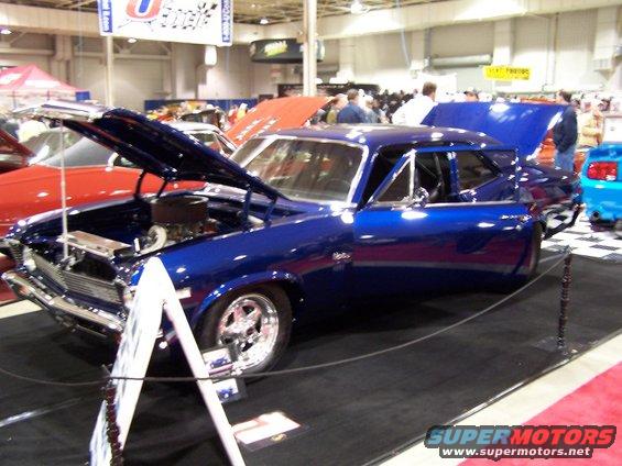 Blue classic car with hood and trunk open at a car show. Other vehicles and people are visible in the background