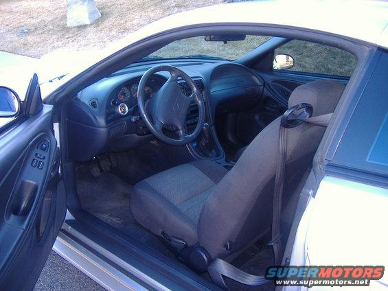 Interior of a white car, showing the driver's seat, dashboard, steering wheel, and door panel. The car has an aftermarket gauge on the dashboard