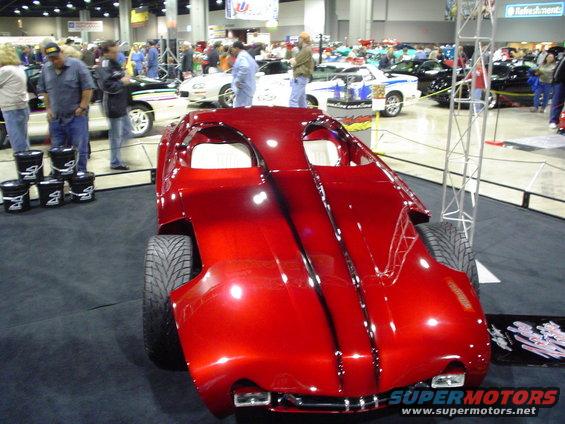 Red custom hot rod with exposed interior and wide rear tires, on display indoors