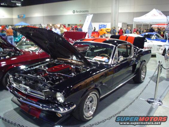 Black 1965 Ford Mustang with orange stripes, hood and trunk open, at a car show