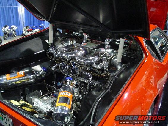Orange car engine bay with chrome intake, two NOS tanks, and motorcycles in background