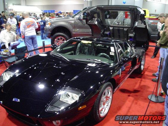 Black Ford GT sports car, 3/4 front view. A modified pickup truck is behind it