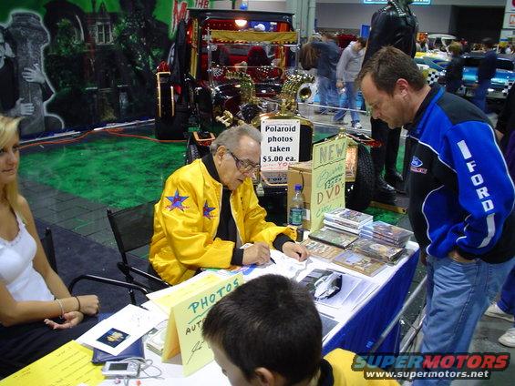 Man in yellow jacket signing autographs at a table with DVDs and photos; another man in a blue jacket stands nearby