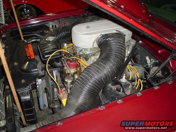 Engine bay of a red vehicle, showing a V8 engine with large air intake hoses. Another red vehicle is visible in the background