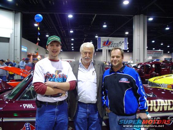 Three men stand in front of custom cars at a show. One car has 'PHIL' and 'HUND' visible on its side