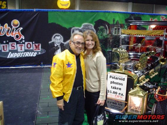Man in yellow jacket and woman pose by a shiny custom car with gold accents and a sign offering Polaroid photos