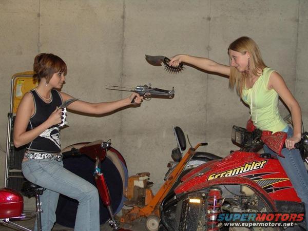 Red ATV with 'Grumbler' text and a woman holding a prop weapon, with another woman on a scooter holding a prop weapon