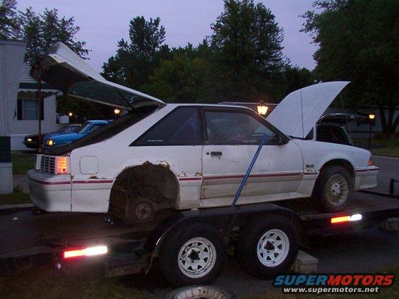 White 1988 Ford Mustang with hood and trunk open, missing rear wheels, on a trailer
