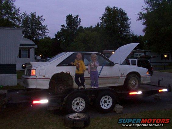 White coupe on trailer with hood up, two kids standing on trailer deck