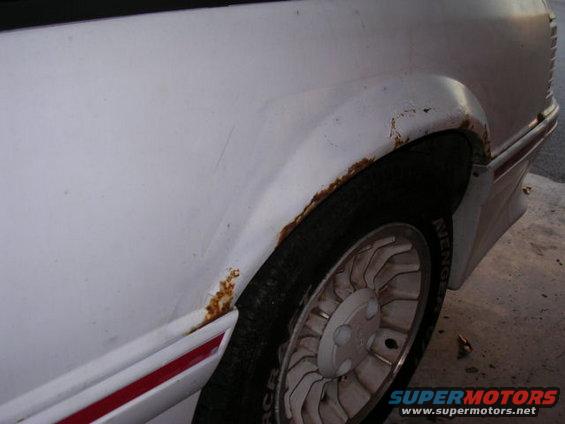 White car's rear wheel well with rust around the fender and a red stripe on the trim