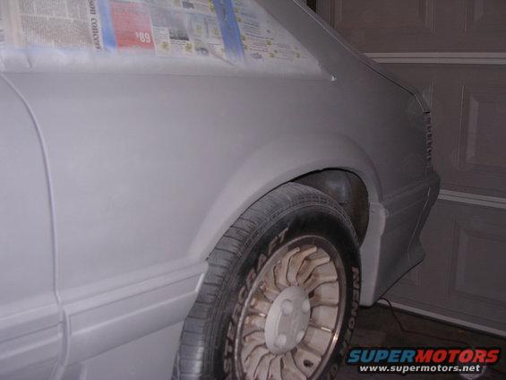 Side view of a car's rear quarter panel and wheel well, covered in primer and masked with newspaper