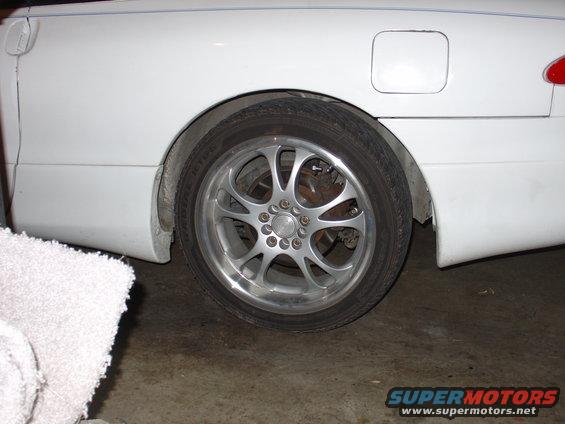 White car's rear wheel with custom rims. Gas cap door visible on the quarter panel