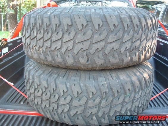Two large off-road tires stacked in a truck bed, secured with red straps. Supermotors.net logo visible