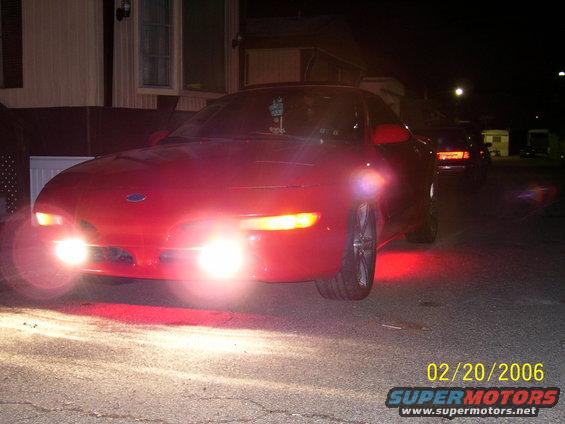 Red coupe with fog lights on, front view. Another car is visible behind