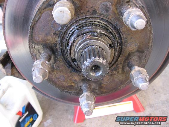 Close-up of a rusty wheel hub assembly, rotor, and lug nuts. A bottle of oil is visible on the left