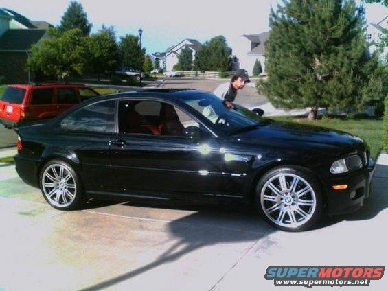 Black BMW coupe parked in driveway, red SUV behind it. Guy leaning on car