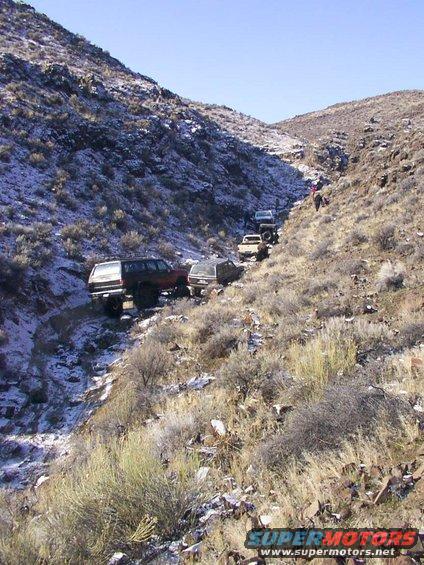 Multiple off-road vehicles driving up a steep, snowy mountain path with sparse vegetation