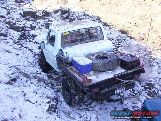 White flatbed truck with spare tire and cooler in the bed, stuck on a snowy, rocky incline