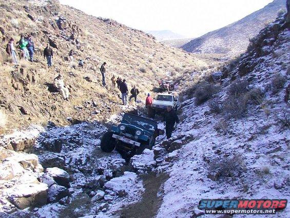 Several off-road vehicles, including a teal Jeep CJ-5, navigating a snowy, rocky trail with people watching