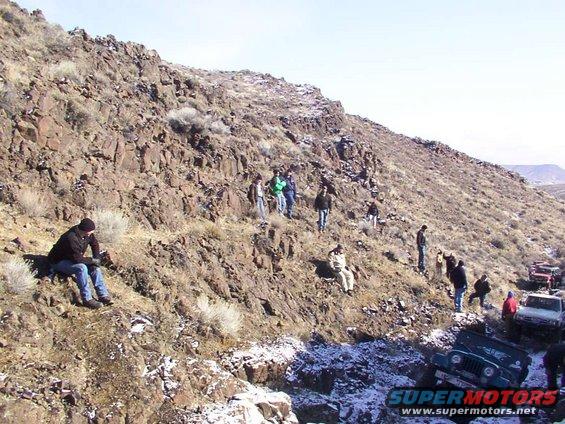 Several off-road vehicles, including a green Jeep, are parked on a snowy, rocky slope with people standing around