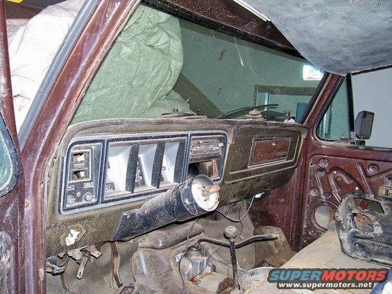 Interior of an old truck, showing the dashboard, steering column, and gear shifter. The dashboard is dusty and some parts are missing