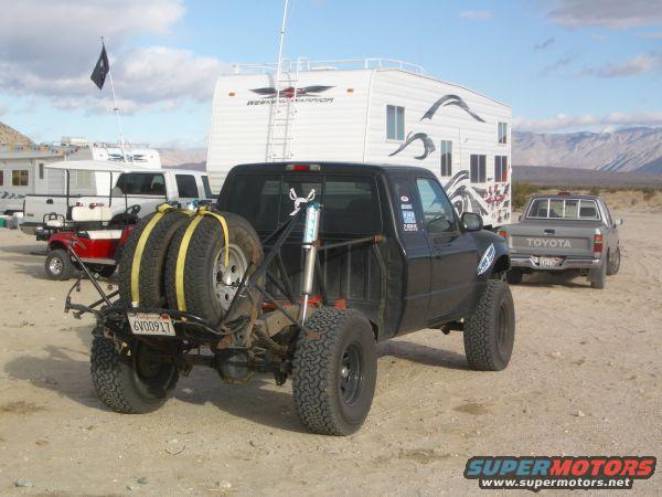 Black pickup truck modified for off-road, with two spare tires mounted on the rear. A silver Toyota truck is parked behind it