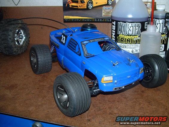 Blue remote control monster truck with large tires, sitting on a brown surface. Bottles of "Monster Horsepower Trinity" fuel are in the background