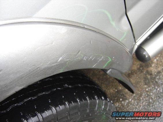 Close-up of a silver car's rear fender showing scratches and green paint marks above the tire