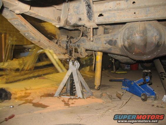 Underside of a vehicle on jack stands, showing the rear axle, leaf springs, and shock absorber. A blue hydraulic jack is also visible